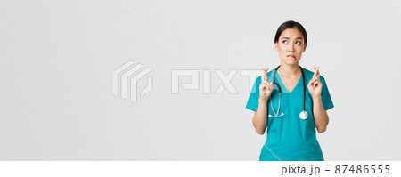 Covid-19, healthcare workers, pandemic concept. Hopeful nervous asian female intern in scrubs waiting for news, doctor cross fingers and biting lip as looking upper left corner, praying 87486555