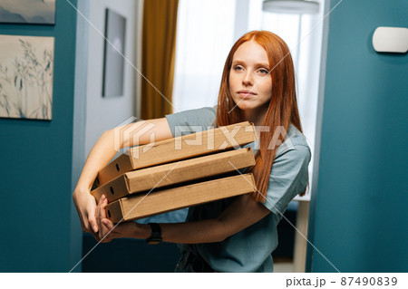 Portrait of pretty young woman holding boxes of hot pizza standing at doorstep to apartment, looking forward. Cute female customer received online order from courier with prepared meal for lunch 87490839