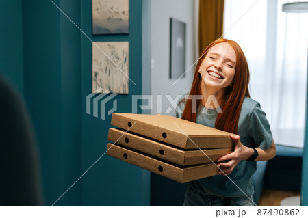 Portrait of excited cheerful young woman holding boxes of hot pizza standing at entrance to apartment. Happy female customer received online order from courier with prepared meal for lunch. 87490862