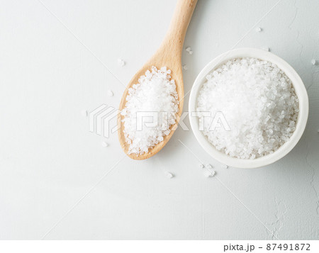 White bowl with large sea salt, wooden spoon, scattered salt crystals on gray stone table. Top view, copy space. 87491872