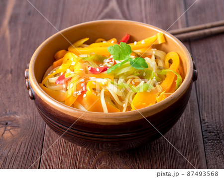 Asian cuisine, noodles stir-fry udon with vegetables. Pepper, carrot, leek, sesame and teriyaki sauce on a brown wood table, side view, close up. Asian cuisine, noodles stir-fry udon with vegetables. Pepper, carrot, leek, sesame and teriyaki sauce on a brown wood table, side view, close up. 87493568