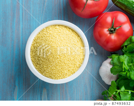 Vegetarian food - wheat, grain of raw couscous in a white bowl on a blue wooden background, fresh vegetables - parsley, cucumbers, tomatoes, top view Vegetarian food - wheat, grain of raw couscous in a white bowl on a blue wooden background, fresh vegetables - parsley, cucumbers, tomatoes, top view 87493632