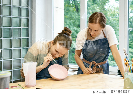 Young skillful pottery master teaching apprentice sculpting and painting mud bowl in art studio Young skillful pottery master teaching apprentice sculpting and painting mud bowl in art studio 87494837