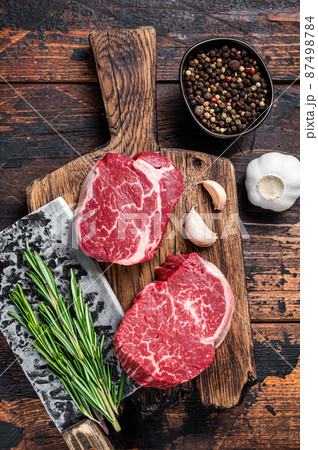 Fresh Raw Fillet Mignon tenderloin steaks on butcher cutting board with meat cleaver. Dark wooden background. Top view Fresh Raw Fillet Mignon tenderloin steaks on butcher cutting board with meat cleaver. Dark wooden background. Top view 87498784