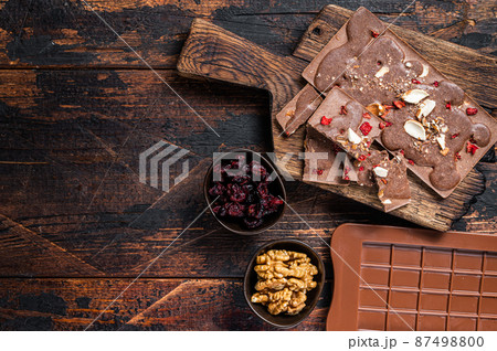 Homemade Craft milk chocolate bar with hazelnuts, peanuts, cranberries and freeze dried raspberries. Dark wooden background. Top view. Copy space 87498800