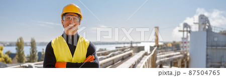 Cheerful male worker standing at industrial site against blue sky Cheerful male worker standing at industrial site against blue sky 87504765