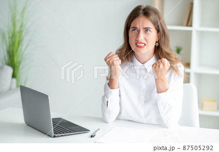 Angry feelings. Aggressive woman. Office worker. Rage lady in white elegant shirt sitting desk opened laptop hands with fists up light room interior. 87505292