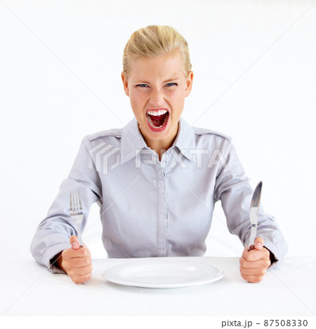 Feed me now. Furious young woman sitting in front of an empty plate while holding her knife and fork. 87508330