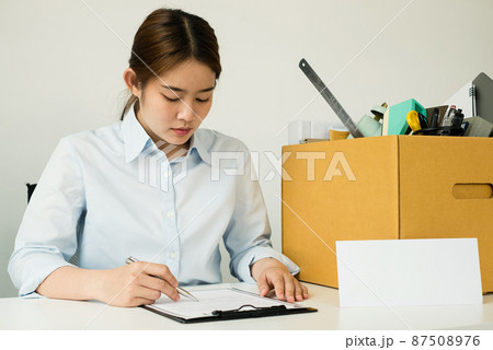 A female office worker, dissatisfied with the termination, packs her belongings in a cardboard box and sits down desperately writing her resignation letter. Concept dismissal and unemployment A female office worker, dissatisfied with the termination, packs her belongings in a cardboard box and sits down desperately writing her resignation letter. Concept dismissal and unemployment 87508976