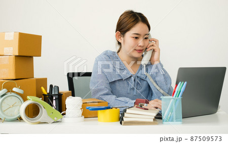 Young Asian woman business owner with many parcel boxes on the table happy online sales job, use the laptop, get an order from customers, take notes, and make arrangements for delivery by post 87509573