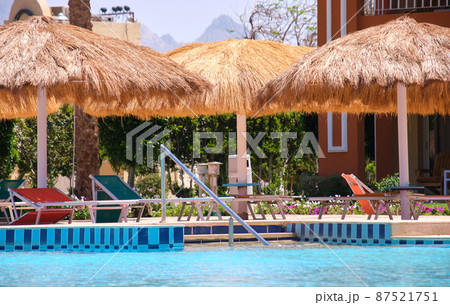 Empty deck chairs under straw shade umbrellas on swimming pool side in tropical resort. Summer vacations and getaway concept Empty deck chairs under straw shade umbrellas on swimming pool side in tropical resort. Summer vacations and getaway concept 87521751