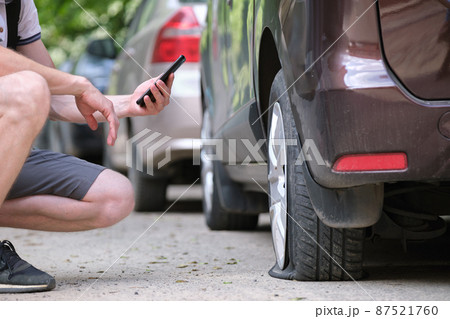Driver calling road service for assistance having vehicle trouble with punctured flat tire on car parked on roadside Driver calling road service for assistance having vehicle trouble with punctured flat tire on car parked on roadside 87521760