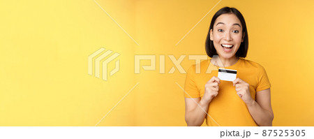 Beautiful asian woman showing credit card and smiling, recommending bank service, standing over yellow background 87525305