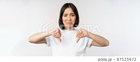 Image of asian woman showing thumbs down, dislike smth, looking disappointed, standing over white background 87525672