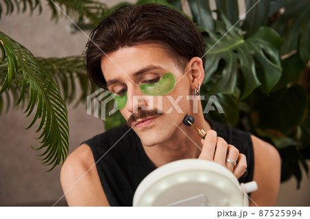 Man putting eye patches on skin and making massage with jade roller while looking at his reflection 87525994