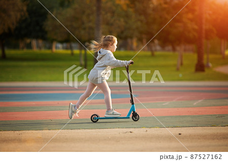 Portrait of active little toddler girl riding scooter on road in park outdoors on summer day. Seasonal child activity sport. Healthy childhood lifestyle Portrait of active little toddler girl riding scooter on road in park outdoors on summer day. Seasonal child activity sport. Healthy childhood lifestyle 87527162