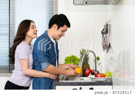 happy couple washing sweet pepper and vegetables in the sink in the kitchen at home. woman is hugging man happy couple washing sweet pepper and vegetables in the sink in the kitchen at home. woman is hugging man 87529317