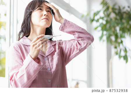 手に持った体温計を見てネガティブな表情になる部屋着の女性 87532938