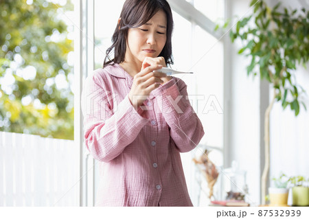 手に持った体温計を見てネガティブな表情になる部屋着の女性 87532939