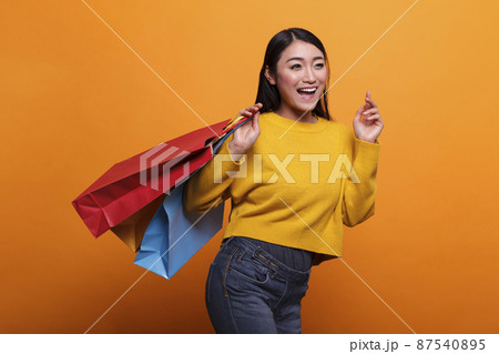 Overjoyed excited beautiful asian woman carrying shopping bags from outlet store while wearing yellow sweater. 87540895