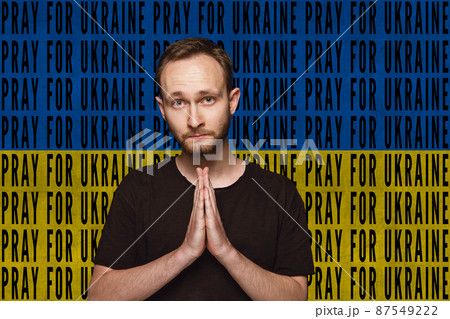 Portrait of Ukrainian man praying for Ukrainian safety isolated over blue and yellow flag background with lettering Pray for Ukraine 87549222