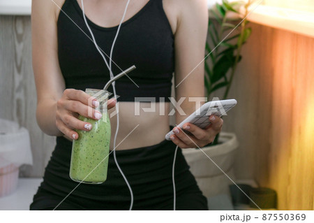 Young millenial woman checking smartphone drinking vegan smoothie drink detox. Woman doing yoga exercises at home. Online tuition Clean eating, weight loss, healthy dieting food concept. Stay healthy 87550369