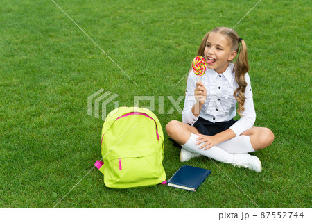 Happy girl child in uniform with school bag and book enjoy eating tasty lollipop sitting on grass Happy girl child in uniform with school bag and book enjoy eating tasty lollipop sitting on grass 87552744