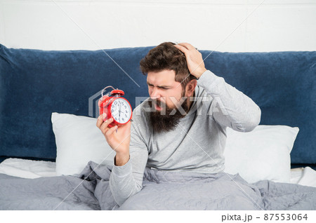 Perplexed guy scratching head holding alarm clock being in bed in morning, oversleep 87553064