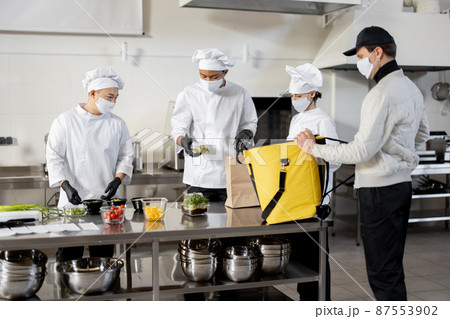 Courier waiting for an order for delivery in the kitchen with chefs preparing takeaway food Courier waiting for an order for delivery in the kitchen with chefs preparing takeaway food 87553902