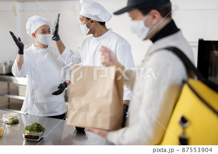 Courier waiting for an order for delivery in the kitchen with chefs preparing takeaway food Courier waiting for an order for delivery in the kitchen with chefs preparing takeaway food 87553904