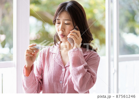 体温計の検温結果を見てどこかに電話をかけるパジャマの女性 87561246