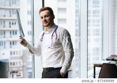 Portrait of handsome male doctor in white uniform examining chest x-ray lungs scan standing near window in hospital office, looking at camera. Concept of medicine and health care. 87568899