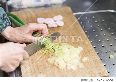 ラーメンの具（ネギとナルト）を包丁で切るシニア女性 87573228