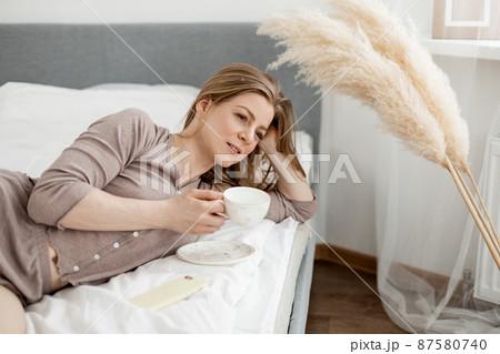 Young beautiful woman in light pajamas enjoys morning in bed with coffee in cozy bedroom. Morning rituals 87580740