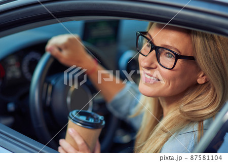 Smiling caucasian businesswoman wearing eyeglasses sitting behind steering wheel of her modern car and buying take away coffee in paper cup 87581104