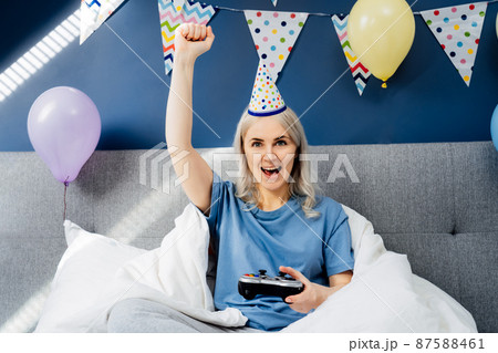 Emotional excited woman in Birthday hat sitting on her cozy bed with controller and playing video game. Birthday party ideas at home. Online time, virtual life, Gaming addiction. Selective focus Emotional excited woman in Birthday hat sitting on her cozy bed with controller and playing video game. Birthday party ideas at home. Online time, virtual life, Gaming addiction. Selective focus 87588461