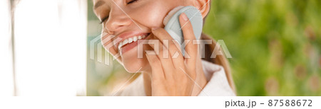 Cropped portrait of young woman wearing bathrobe smiling while using silicone face brush to cleanse skin 87588672