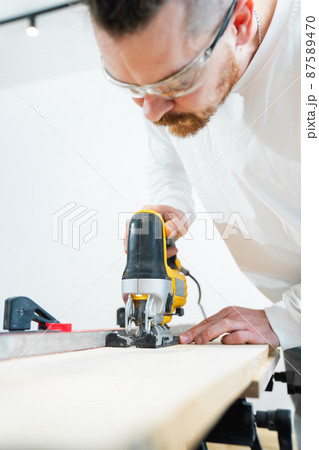 carpenter in protective gloves and glasses cuts wood with electric jigsaw carpenter in protective gloves and glasses cuts wood with electric jigsaw 87589470