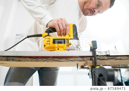 carpenter in protective gloves and glasses cuts wood with electric jigsaw 87589471
