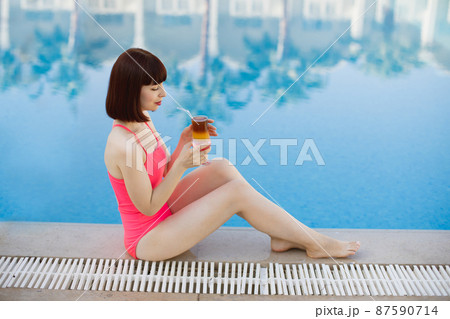 Pretty lady with alcohol drink sitting near the blue pool 87590714