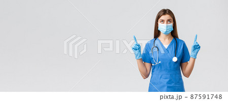 Covid-19, preventing virus, health, healthcare workers and quarantine concept. Curious female doctor or nurse in blue scrubs, medical mask reading banner, pointing fingers up Covid-19, preventing virus, health, healthcare workers and quarantine concept. Curious female doctor or nurse in blue scrubs, medical mask reading banner, pointing fingers up 87591748