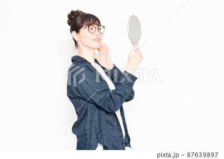 白い背景の前に立って手鏡で自分の肌を見る眼鏡をかけている女性 87639897