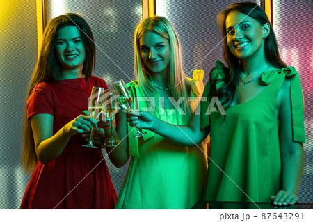 Happy girls clinking glasses of sparkling wine in colorful lighting of nightclub Happy girls clinking glasses of sparkling wine in colorful lighting of nightclub 87643291