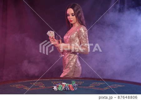 Woman in golden dress showing cards, holding champagne while posing at playing table in casino. Black, smoke background. Gambling, poker. Close-up. Woman in golden dress showing cards, holding champagne while posing at playing table in casino. Black, smoke background. Gambling, poker. Close-up. 87648806