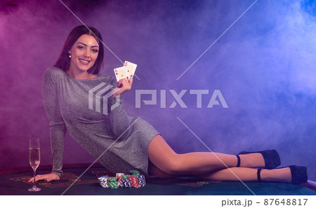 Girl in gray dress playing poker at casino, holding cards, laying on table with chips and champagne on it. Black, smoke background. Close-up. Girl in gray dress playing poker at casino, holding cards, laying on table with chips and champagne on it. Black, smoke background. Close-up. 87648817