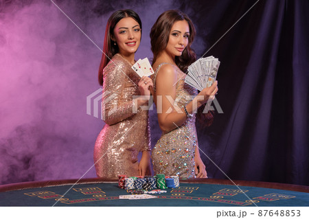 Two women showing cards and money, posing at playing table in casino. Black, smoke background with colorful backlights. Gambling, poker. Close-up. Two women showing cards and money, posing at playing table in casino. Black, smoke background with colorful backlights. Gambling, poker. Close-up. 87648853