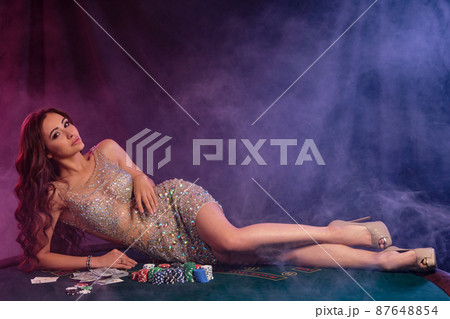 Girl in golden dress playing poker at casino, laying on table with chips, money, cards on it. Black, smoke background. Gambling. Close-up. Girl in golden dress playing poker at casino, laying on table with chips, money, cards on it. Black, smoke background. Gambling. Close-up. 87648854