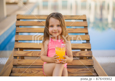 Portrait of little cute girl in pink swimsuit with orange juice glass resting on a wooden lounge. Portrait of little cute girl in pink swimsuit with orange juice glass resting on a wooden lounge. 87649902