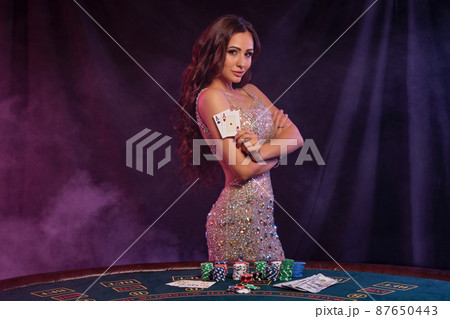 Girl in golden dress playing poker at casino, holding two aces. Posing at table with chips, money on it. Black, smoke background. Close-up. Girl in golden dress playing poker at casino, holding two aces. Posing at table with chips, money on it. Black, smoke background. Close-up. 87650443