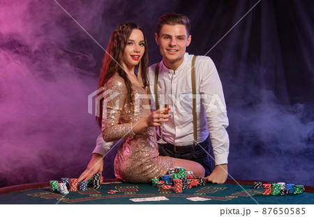 Man and woman playing poker at casino, celebrating win at table with stacks of chips, cards, champagne. Black, smoke background. Close-up. 87650585
